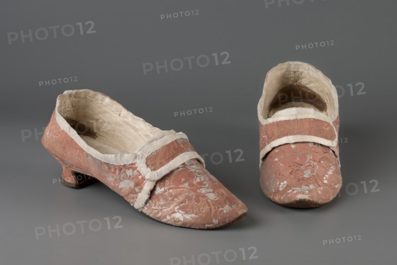 A pair of women's slippers made of patterned silk damask lined with leather. Dark pink outer fabric with a white floral motif. Shallow slippers, fitted with two decorative straps at the front. 3.5 cm heel, tapering down from mid-height. Symmetrical soles, pointed towards the front. Fabric-covered heel. Hand-sewn. Toes lined with canvas; heel lined with leather. Endpapers made of thin white leather. 2nd half of the 18th century, Europe. Height 9 cm, length 25 cm., A pair of low-heeled shoes sit on a neutral gray background, both covered in a faded pink fabric with a floral woven pattern in a lighter color; the shoes have pale off-white trim around the openings and across the insteps, and the interiors show a worn beige lining; one shoe is shown from an angled side view revealing a short curved heel and scuffed sole, while the other is shown from the front with a rounded toe and visible interior shadowing.