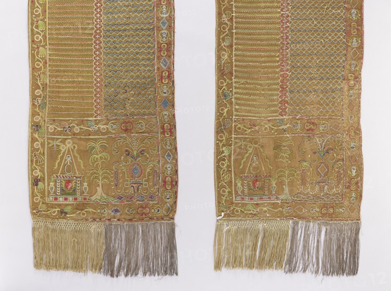 Kontusz belt, carp scale ornament, one-sided belts, plant ornaments, Polish belts, Polish costumes, kontusz belts, belts, fashion accessories, Poland culture, clothing accessories, men's clothing accessories. At the heads of the belt is a palm tree in the center; on the sides, on one side is a vase of flowers, on the other is an arbor with a heart in the center. Frame of plant twigs on one side, geometric ornament on the other. The belt has carp scales on one side and transverse stripes of plant twigs on the other. Background color: olive; ornament colors: blue, yellow, red, and green. Fringe silver on one side, beige on the other. 18th century, Poland. Length 388.5 cm, width 45.5 cm., The image shows a pair of long, narrow textile panels laid side by side on a pale background, each panel in a warm golden-brown fabric embroidered with intricate decorative motifs in colors including gold, green, red, blue, pink, and white; both panels have a wide embroidered border and internal patterne...