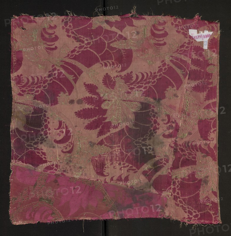 Fabric fragment, silk, two-tone silk damask, patterned with plant motifs in the Bizarre style, ca. 1712, probably Italy. Sub-backing for the brocaded decoration is a drawing highlighted by a lighter weft color and simple weave against a darker cherry warp-covered satin background. Brocaded decoration uses gold braided thread bound with a weft weave; metal thread spun in pairs in regular shifts, without knotting longest with over 20 warps. The lighter sub-backing consists of large plant-like forms winding opposite the golden decoration, resembling palmettes suspended on a slender twig. Decoration: slender, sinuously winding golden branches bearing fanciful flowers surrounding light pink forms resembling crustacean shells and palmettes composed of leaves with jagged edges. Colors: cherry background, light pink subground, gold stitching. Weave: background – warp-covered satin cherry warp; pattern – plain weave with a grosgrain effect yellow weft; damask subground – warp-covered satin cher...