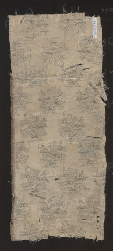Fabric fragment, 17th century, France. Fragment of silk patterned fabric with preserved design: leafy twigs tied with a ribbon, arranged in rows with shifts and changes of direction. Colors: white and silver background, silver motifs. Pattern size: 12 x 8.7 cm. Overall dimensions: height 46.5 cm, width 39 cm., A rectangular piece of worn fabric with frayed edges placed on a dark background, showing a repeating floral-like woven pattern in muted gray against a beige ground; the fabric has several small tears and holes, loose threads, and patches of wear, with faint darker smudges and slight discoloration in places, and a narrow strip of folded or layered fabric near the top right edge.