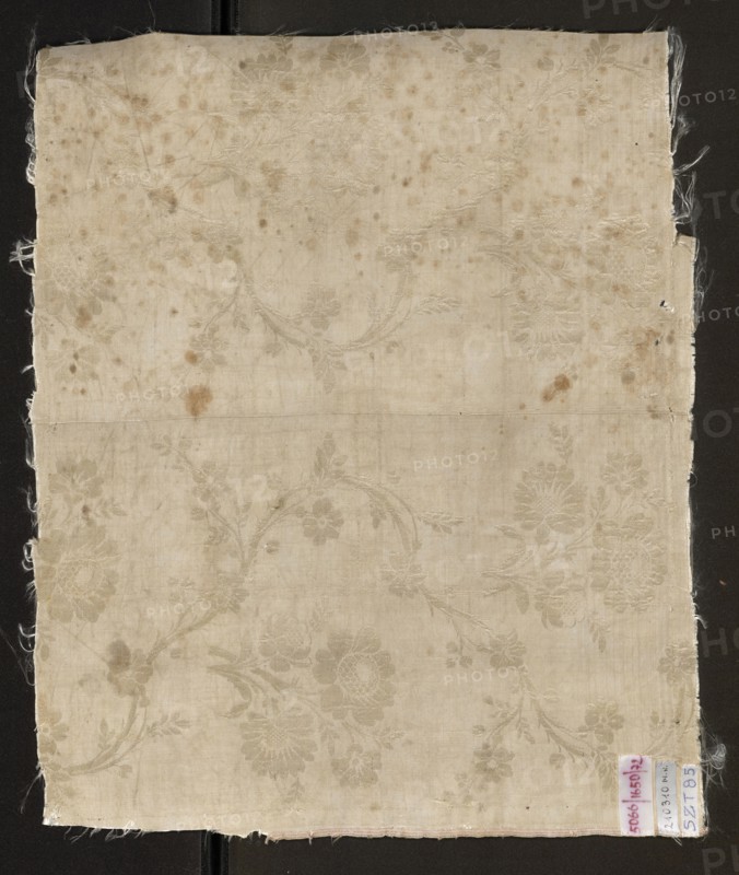 Fabric fragment, silk, patterned floral double-warp fabric. Cream and silver lamé with a pattern of twisting floral branches. 18th century, France; height 47 cm, width 39 cm., A square piece of fabric with frayed edges shown against a dark background, the fabric is an off-white to beige color with a subtle floral damask pattern visible across its surface; there are several brownish stains and spots scattered on the fabric and a faint horizontal fold line across the middle; a small rectangular tag is attached near one corner with a hint of pink and blue visible on it.