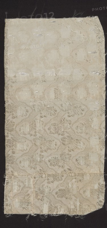 Fragment of fabric, fabrics, fragments of a larger textile object, silk fabric, 18th-century silk fabric, patterned fabric: droguet maurois sur fond taffeta, à petits dessins liserés. A similar fragment constitutes a fragment of a men's habit inventory no. 33068. Report: 5.5 x 7 cm. A drawing of a similar pattern appears in L'Art du fabricant d'étoffes de soie by J. Paulet fig. 195. 1760, Sweden; fabrics, height 38 cm, width 19 cm., A rectangular piece of fabric with frayed edges lies against a dark background; the fabric is a pale off-white or cream color with a subtle woven pattern forming repeating rounded shapes and floral-like motifs in slightly darker and lighter shades of cream; the surface shows a few horizontal slubs and irregularities where threads are worn or pulled, and there are several areas with faint discoloration and small loose fibers along the borders.