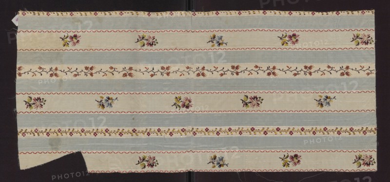 Fabric fragment, fragment of silk fabric with a pattern of alternating vertical stripes of blue and cream; colorful motifs of small flower sprigs and tiny floral tendrils on the light stripes. 19th century, France. Silk. Height 72 cm, width 32.5 cm., A rectangular piece of fabric with alternating horizontal bands in pale blue and off-white, decorated with a repeating floral motif; the off-white bands contain small embroidered bouquets in colors including pink, yellow, green, and blue, separated by wavy red trim, while a few narrow off-white strips feature a garland of small flowers in shades of rust, brown, and muted red on a dark branch; there is a narrow decorative border with tiny floral clusters in red and yellow against an off-white ground, and the fabric edges show some fraying and a missing corner on one side, all set against a dark background.