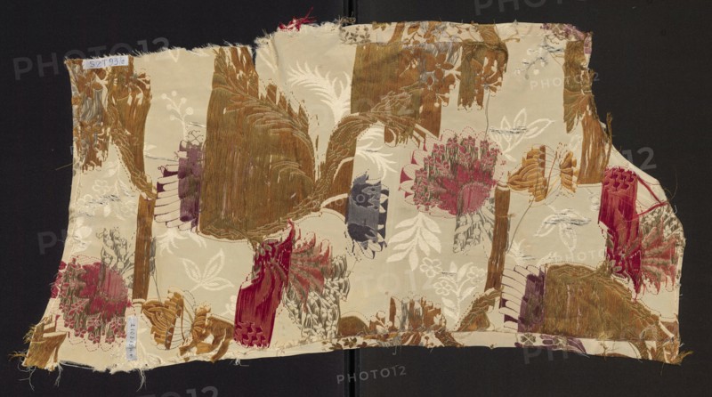 Fragment of upholstery fabric, silk and cotton brocade with cream grosgrain plain-weave background, brocaded in colorful threads and metal threads. Floral and fantastic motifs: winding twigs with fanciful flowers and plumes of leaves resembling bird wings; patterned fabric with preserved selvedge. Colors: red, dark and light pink, dark and light brown, damp green, blue, silver metal thread, copper metal thread. Dimensions: height approx. 64–71 cm, width 32 cm; edge width 0.9 cm. Possibly French, 18th century ca. 1760–1777 or early 19th century influence., A rectangular piece of fabric with frayed edges laying on a dark background, showing a patterned surface with floral and abstract woven motifs in shades of cream, beige, brown, gold, rust, red, pink, purple, gray, and green; several clustered flower-like shapes and leaf forms are woven into the cloth, along with textured vertical and curved bands that appear distressed or worn in places, and a few loose threads visible around the bord...