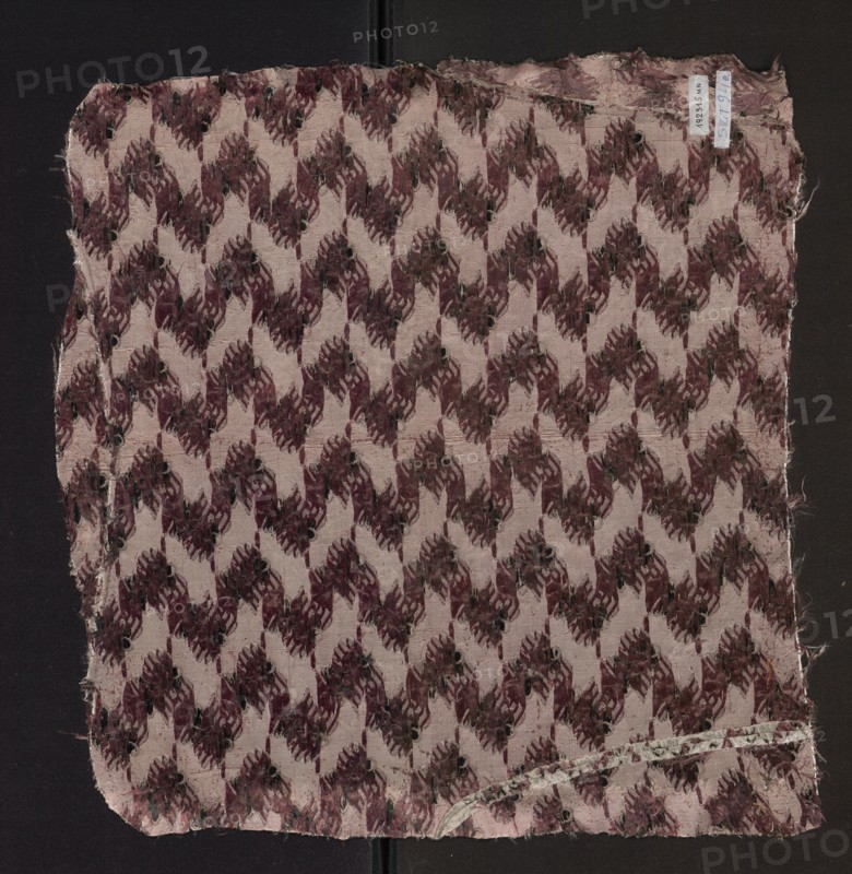 Fabric fragment of patterned silk with floral motifs on a white background motifs in pale pink, plum, and black. 2nd half of the 18th century, France. Height 51 cm, width 48 cm., A square piece of frayed fabric with a repeating geometric floral-like pattern in shades of mauve, burgundy, dark brown, and black on a pale pink background, laid against a dark surface; the fabric edges are worn and uneven with some loose threads and a narrow folded hem along one edge, and a small curved strip of lighter material with a faint spotted design is visible near one corner.