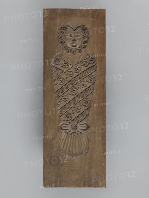Double-sided gingerbread mould. Side A: image of a bachelor; Side B: baby in swaddling clothes., woodcarver or gingerbread maker. Double-sided rectangular mould. One side shows a man in a long wig and a tricorn hat, dressed in a costume probably dating from the turn of the 17th and 18th centuries: justaucorps with decorative cuffs, culottes, stockings, and high-heeled shoes; at the figure's feet are decorative motifs of flowers and geometric elements and the date 1827. The other side depicts a baby in swaddling clothes richly decorated with geometric and plant ornament. On the edge is an inscription: FW FIEBIGER. a. BAUTZEN. 17 signature of the manufacturer and the beginning of the date., wood gingerbread mould, folk motifs, plant ornaments, floral decorations, historical costumes; Silesia Prussia; 1742–1918 / Bautzen. Date: 1827. Function: form. Dimensions: height 64 cm, width 22.5 cm, depth 4.3 cm., A carved wooden panel shows a stylized wrapped figure with a rounded head and visible...