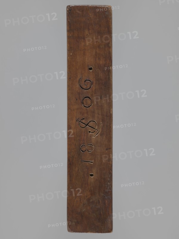 Double-sided gingerbread mould. Side A: bison. Side B: three figures — a bachelor in early 19th-century costume hat, justokor, artificial trousers, high boots, a baby in swaddling clothes decorated with a plant twig, and a fish with an elongated, scaled body. Rectangular wooden mould with the date 1806 engraved on the edge; the date includes the letter S over an anchor manufacturer or gingerbread maker's mark. Gingerbread mould; folk art/folk customs; zoomorphic and figurative motifs; Striding Bull iconography. Dated 1806. Province of Silesia Prussia; 1742–1918. Form. Dimensions: height 19.5 cm, width 10.5 cm, depth 4.3 cm., woodcarver or gingerbread maker., A long vertical wooden plank with a smooth, worn surface in brown tones against a plain light gray background; the plank shows a few carved grooves and shallow round holes along its center, areas of darker and lighter brown from wear, and some subtle scratches and dents.