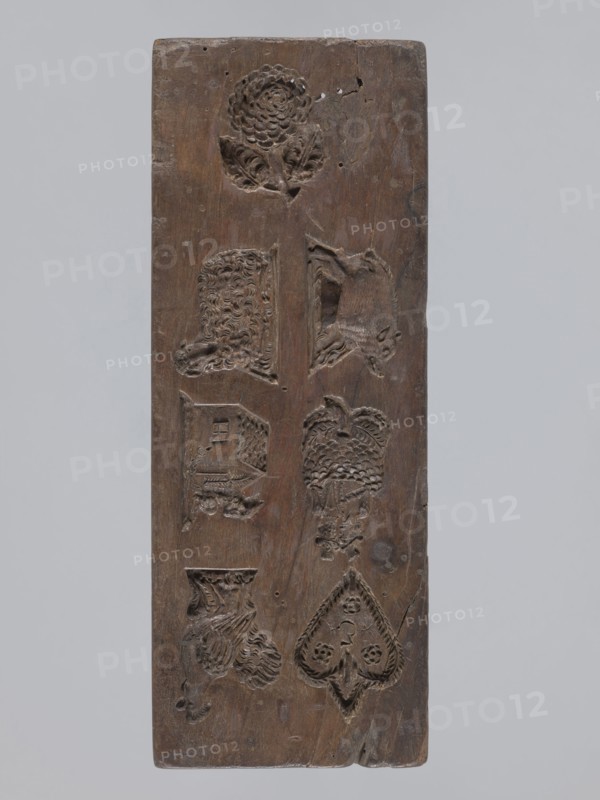 Double-sided gingerbread mold; obverse: seven different motifs — a heart, a rooster, a dog in front of a kennel, a woman and a man in costumes from the second half of the 19th century, a wild boar, a lamb and a fish; reverse: a fish covered with scales. 19th century, Province of Silesia 1742–1918. Height 28.5 cm, width 10.7 cm, depth 5.5 cm., A rectangular wooden board with a vertical orientation showing carved recessed motifs arranged in two vertical columns; the wood is brown with variations in tone and a weathered, slightly worn surface; a carved flower with layered petals and leaves appears near the top, and below it are carved animal and object shapes including a textured sheep-like head, a reclining animal, a house-like form, a bird with patterned feathers, a draped textile or flowing mane, and a heart-shaped spade motif with decorative inner carving; the carvings show tool marks and varying depths, and the board has small holes, surface cracks, and areas of darker brown patina.