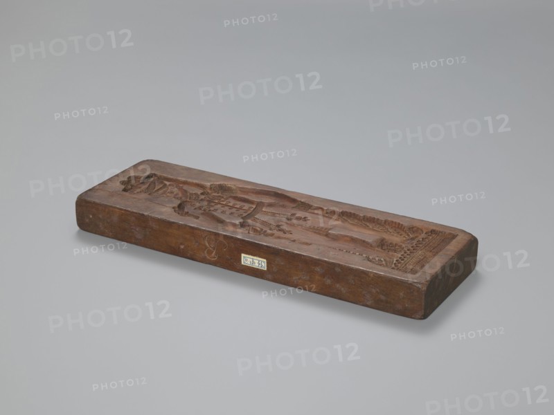 Double-sided gingerbread mould. Side A: image of a soldier; Side B: baby in swaddling clothes., woodcarver or gingerbread maker. Gingerbread mould, unknown origin. Decorative art; male figures; historical costumes; gingerbread; plant ornaments; military uniforms; folk customs; Silesia Prussia; 1742–1918; folk motifs. Double-sided, rectangular mould: one side shows a soldier in a parade uniform from the beginning of the 19th century, wearing a high shako with a tassel, epaulettes and decorative sleeves, an order pinned on the chest, a sabre with decorative tassel at the leg; geometric and plant decoration at the bottom. The other side shows a baby in swaddling clothes richly decorated with geometric and plant ornament. On the edge, the letter engraved B initial of the woodcarver or gingerbread maker. 19th century, Silesia Province 1742–1918. Form; height 35.3 cm, width 11.6 cm, depth 3.5 cm., A rectangular wooden block rests on a smooth grey surface, showing a carved shallow relief of a...
