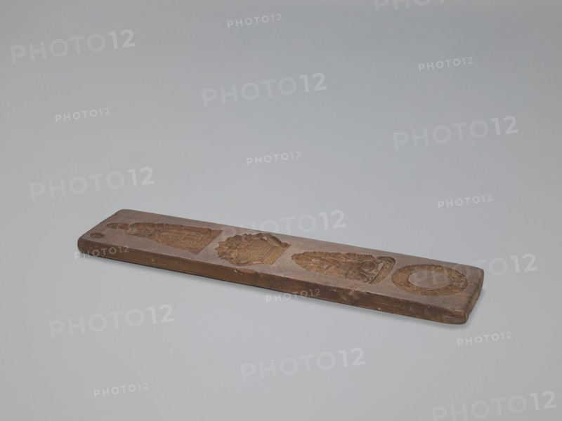 Double-sided gingerbread mould. Obverse: four motifs — at the top a female figure in a long patterned dress with a low neckline; below, a basket of flowers; below that, a seated young naked man with long hair sitting among flowers and smoking a pipe; at the feet of the figure a skull with crossbones and the inscription MEMENTO MORI; at the very bottom a wreath woven from leaves and flowers. Reverse: five images of infants in patterned swaddling clothes. Materials/subject: gingerbread mould, gingerbread, decorative art; motifs: wreaths, female figures in rich dresses, Transience personification, floral decorations, allegorical representations, infants, folk motifs. Double-sided, rectangular mould. 19th century. Province of Silesia 1742–1918. Dimensions: height 36.5 cm; width 8.8 cm; depth 1.5 cm. /origin., A long rectangular wooden board rests on a light gray surface, showing a medium brown wood tone with darker brown carved recessed shapes along its length; the board has worn edges and...