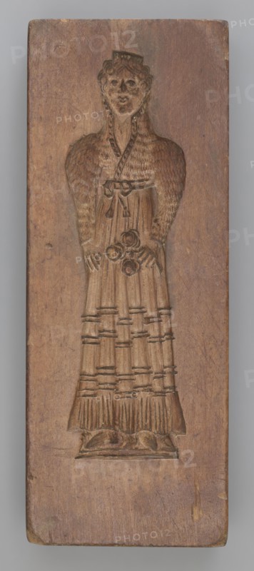 Double-sided gingerbread mould. Side A: female figure in a decorative, pleated early 19th-century dress, holding a bouquet of roses. Side B: stylised fish covered with scales shown in profile. Rectangular wooden mould, double-sided. Edge engraved with the date 1808 and, centered in the date, the letter S against an anchor mark of the woodcarver/gingerbread maker—owner of the form. Materials: wood; function: gingerbread mould; motifs: fish, flowers, female figures in rich historical costume, zoomorphic and folk motifs. Maker: unknown woodcarver or gingerbread maker. Origin: Silesia Province of Silesia, dated 1808. Dimensions: height 19.8 cm, width 7.3 cm, depth 4 cm., A carved wooden panel shows a full-length relief of a standing figure with a rounded head and stylized facial features, curly hair, and a small headpiece. The figure wears a long, detailed dress with a textured upper section suggesting a patterned or feathered texture and a skirt with vertical pleats and horizontal bands. ...