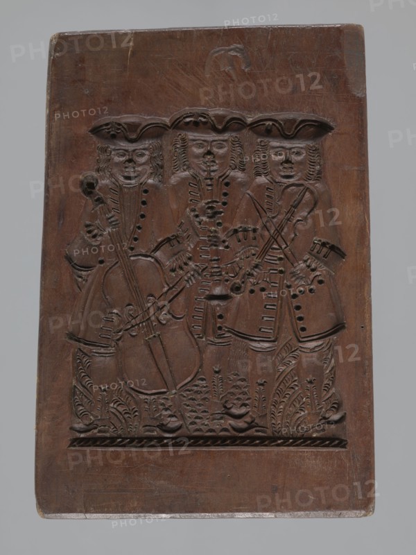 Double-sided gingerbread mould. Obverse: group of musicians; Reverse: three fish. Double-sided, rectangular form; on one side a group of three musicians in costumes typical of the 17th–18th centuries—hat, justaucorps, culottes and stockings—the first playing a double bass, the second a trumpet and the third a violin; floral and plant decoration at the bottom. Reverse: three stylized elongated fish covered with scales, shown in profile and arranged in a row. Gingerbread mould; decorative art; folk motifs; zoomorphic motifs; floral decorations. Early 19th century, Province of Silesia 1742–1918. Height 21.5 cm, width 15 cm, depth 2.8 cm., A rectangular wooden panel with a carved scene of a group of three standing figures wearing tricorn-style hats and long coats, each figure holding a stringed instrument and shown with detailed facial features and curled hair; the carving includes decorative carved foliage at the figures' feet and patterned borders near the bottom. The wood is a medium to...