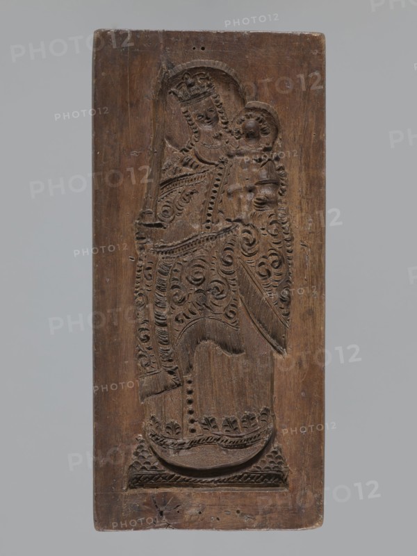 Double-sided gingerbread mould. Obverse: image of Madonna and Child; reverse: small image of Madonna in an oval. Double-sided, rectangular form; on one side a figure of Madonna and Child, with a crown on her head, in a long patterned robe decorated with various geometric and plant motifs; the Madonna holds the Child in her left hand and a sword in her right. The other side: a smaller figure of the Madonna in an oval, surrounded by rays and a laurel wreath. Materials/function: gingerbread mould; style: decorative folk/religious motifs. Iconography: Madonna and Child, crown, sword, wreaths. Date: 19th century. Place: Silesia Province of Silesia, 1742–1918. Dimensions: height 25.8 cm, width 12 cm, depth 4.5 cm., A rectangular wooden panel carved in brown shows a raised relief of a robed figure holding a smaller robed figure; the larger figure wears a crown and both figures have stylized curled hair and carved halo shapes behind their heads. The robes and surrounding areas feature carved s...