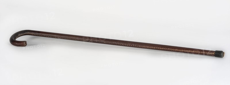 Walking stick, maker, wooden walking stick covered with brown leather along its entire length, rubber-coated foot; 20th century, Europe; men's fashion accessory; length 81 cm, width 7 cm., A single curved walking stick lies on a light background, showing a hooked handle and a straight shaft; the stick is primarily dark brown with lighter tan stitching or decorative inlay running along its length, the hooked end is rounded and the opposite tip is capped with a short, darker piece.