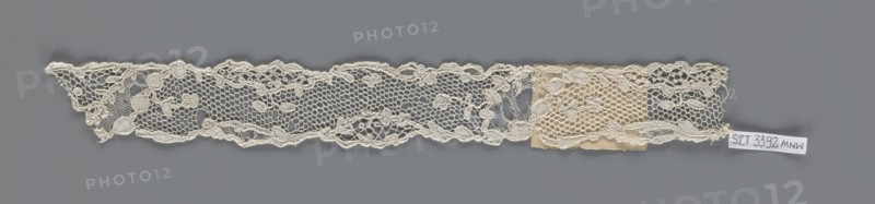 Lace fragment, lace with an ornament carried on two edges by a free tendril and carried diagonally through the central part, creating large fields filled with a net of large knitted meshes; mid-18th century, France; fabric; width 3.5 cm, length 28 cm., A long narrow strip of lace placed against a gray background, showing a delicate openwork pattern with a scalloped top edge and an irregular bottom edge; the lace features a honeycomb-like mesh interspersed with floral and leaf motifs and raised corded outlines, and a small patch of slightly darker fabric is attached near one end; visible colors include off-white and cream in the lace and patch, and gray in the background.