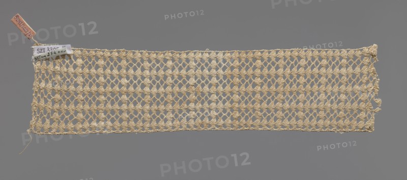 Fragment of crocheted lace — section of a belt, cream-colored, with motifs of semicircular shells and small rhombuses in rows. Lace fragment, width 7 cm, length 31 cm. 1st half of the 19th century, Germany ., A rectangular piece of openwork fabric with a netlike pattern made from a beige yarn stretches horizontally against a solid gray background, showing a repeating diamond-shaped lattice with textured clusters at the intersections and some loose threads at the edges; a small fabric tag and a narrow leather or tan-colored tag are attached at the upper left corner.