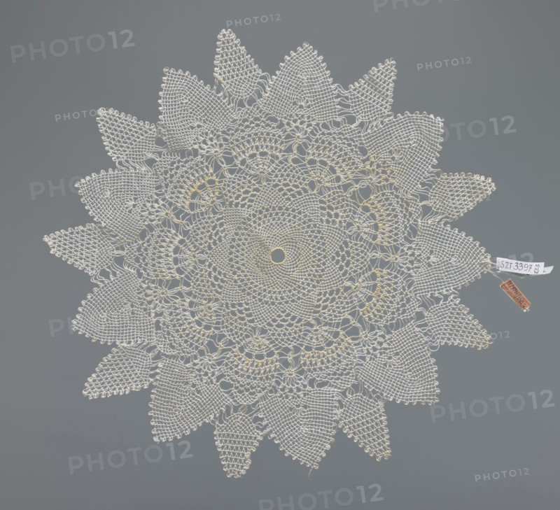 Napkin, napkin, lace napkin, white, round with large triangular teeth along the edge, concentric and radial geometric ornament, 20th century, Poland, fabric, lace, diameter 33 cm, A round crocheted doily with a lacy floral pattern in off-white and pale cream tones laid flat against a solid medium-gray background, featuring a central circular motif surrounded by concentric rings of openwork and petal-like shapes, with a scalloped outer edge of pointed leaf motifs; a small fabric tag and a small rectangular tag in brown are attached near the edge.