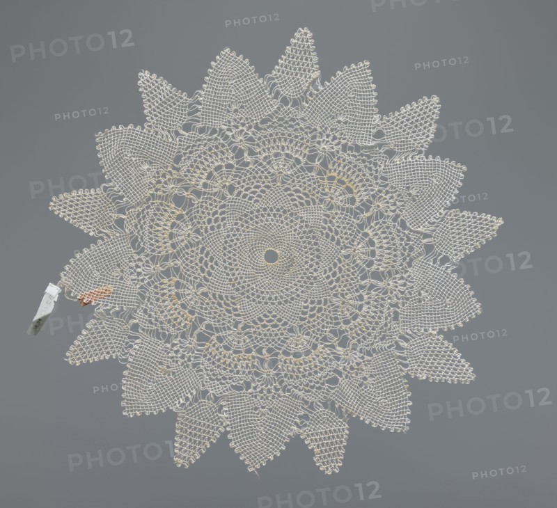 Napkin, napkin, lace, lace napkin, white, round with large triangular teeth along the edge; concentric and radial geometric ornament; 20th century, Poland; fabric, lace; diameter 33 cm, A circular, lace-like doily with an intricate radial pattern forming petal-shaped points around the edge, displayed flat against a smooth gray background; the doily is primarily off-white with subtle hints of pale yellow woven into its center and some inner rings, and there is a small clothespin-like clip attached near the left edge that is light gray with a touch of reddish-brown material nearby.