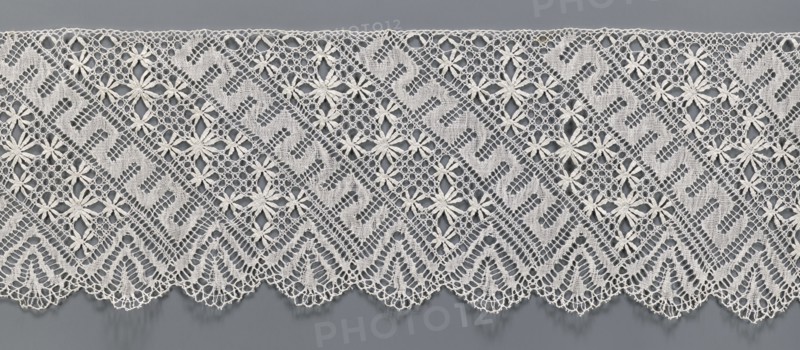 Lace fragment, bobbin lace. White, section of a strip with a pattern of diagonal stripes with rosettes and a meander; one edge serrated. 19th century, Europe. Width 17.5 cm, length 395 cm., A length of lace with an open, geometric floral pattern lies against a smooth background. The lace features repeating diagonal bands of mesh and solid woven sections forming stylized shapes, interspersed with daisy-like floral motifs with petal clusters. The lower edge of the lace has a scalloped border with triangular, fan-like motifs and delicate looped trim. Visible colors include off-white or cream for the lace and a medium gray background.