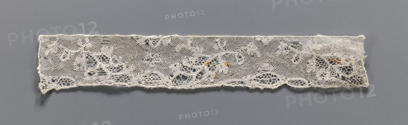 Lace fragment, bobbin lace Valenciennes, white, continuous floral pattern with Rococo motifs, 18th century, width 4 cm, length 23 cm, A horizontal strip of delicate lace with a floral and leaf pattern laid against a smooth background; the lace is off-white with subtle variations in tone and a few small areas of brownish discoloration, showing open mesh sections and more densely stitched floral motifs, scalloped lower edge, and irregular frayed ends, all set on a flat medium gray backdrop.