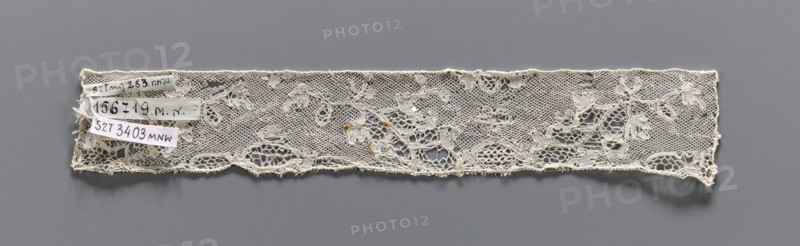 Lace fragment, bobbin lace, Valenciennes lace. Fragment of white bobbin lace with a continuous floral pattern and Rococo motifs. 18th century. Width 4 cm, length 23 cm., A narrow strip of lace with a floral and netted pattern lies horizontally against a solid background, showing a delicate open weave with floral motifs and scalloped lower edge; the lace appears off-white to cream with a few small darker discolorations, and the background is a uniform gray.
