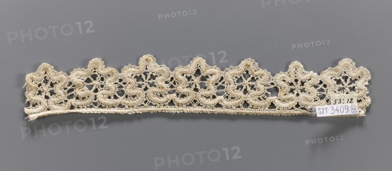 Lace fragment, needle-stitched lace, upper edge straight, lower edge serrated to match the motifs. The pattern of the motifs and the upper edge are emphasized by thickly sewn threads. 19th century, Europe. Height 4 cm, width 27 cm., A narrow length of intricately patterned lace in an off-white or cream color is shown laid horizontally against a flat gray background; the lace has a scalloped top edge with a repeating floral-like motif formed by openwork loops and dense embroidered areas, and a straight lower edge with a fine woven border; there are small areas of slightly darker discoloration within the lace and a small paper tag attached near one end that contrasts with white and light tones.