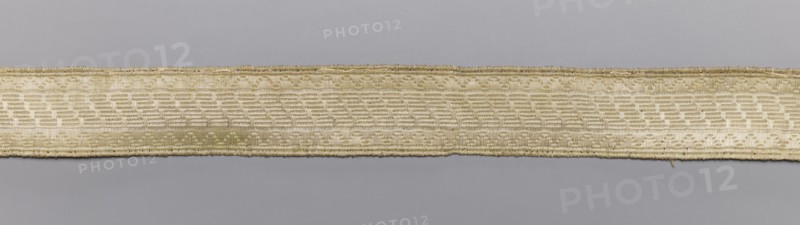 Gallon, galloons haberdashery, geometric ornaments, white warp, weft - wire - drawn thread. Faint ornament, broken line at the edge, geometric ornament in the center: warp stripes interrupted by diagonal, stepped lines. 19th century, Europe. Fabric haberdashery, width 2.2 cm, length 77 cm., A horizontal woven band runs across the image against a plain background, showing a textured pattern of interlaced threads and a slightly raised border along its top and bottom edges; the band features a repeating rectangular weave and some irregularities in thread thickness, with loose fiber ends visible at one point, and the background appears smooth and even. The visible colors are beige, cream, light tan, and gray.
