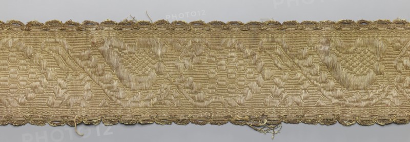 Gallon, gallons haberdashery, origin unknown, plant ornaments, 19th century, Lithuania, fabric, haberdashery, width 9 cm, length 57 cm, A horizontal strip of woven fabric with a repeating geometric pattern in shades of gold and beige, showing a raised, textured weave and subtle sheen; the top and bottom edges have a scalloped decorative border in metallic gold threads with some loose fibers and fraying; the central field displays repeating triangular and circular motifs created by variations in weave height and direction, giving a mix of matte and shiny surfaces in gold, beige, and light tan tones.