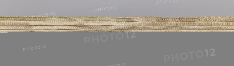 Gallon, haberdashery; geometric ornaments; 19th century, Europe; fabric; width 3 cm, length 54 cm; used as a haberdashery trim., A horizontal scene dominated by a long, narrow woven band with visible texture and layered ridges, showing natural beige and tan fibers with subtle greenish and light brown accents along the upper edge, set against a smooth pale gray background above and a solid mid-gray area below.
