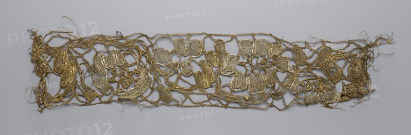 Lace fragment, lace, 2nd half of the 18th century, possibly Flanders, fabric, width 7.5 cm, length 35 cm., A long, narrow, irregularly shaped openwork piece made of metallic thread in gold tones laid flat against a smooth light gray background, with tangled loops and braided strands forming a loose network and a few denser woven patches, frayed loose ends and wispy threads extending from the edges, and subtle variations in the gold appearance where the threads catch the light.