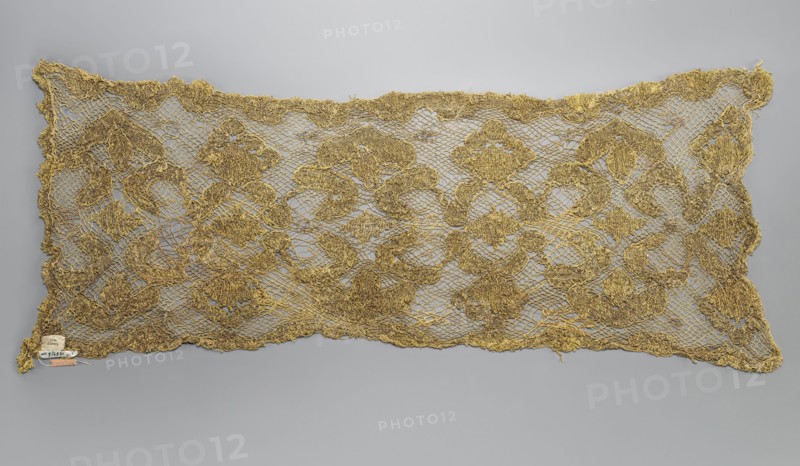 Fragment of lace, lace, 17th/18th century, Belgium, fabric, lace, width 31 cm, length 76 cm, A rectangular piece of openwork fabric laid flat against a neutral background, showing a repeating ornate floral and scroll-like pattern made from metallic threads in shades of gold and bronze, with the ground formed by a net-like mesh of thin threads and the decorative motifs appearing raised and textured; the edges are irregular and scalloped, and a small label-like object is attached near one corner.