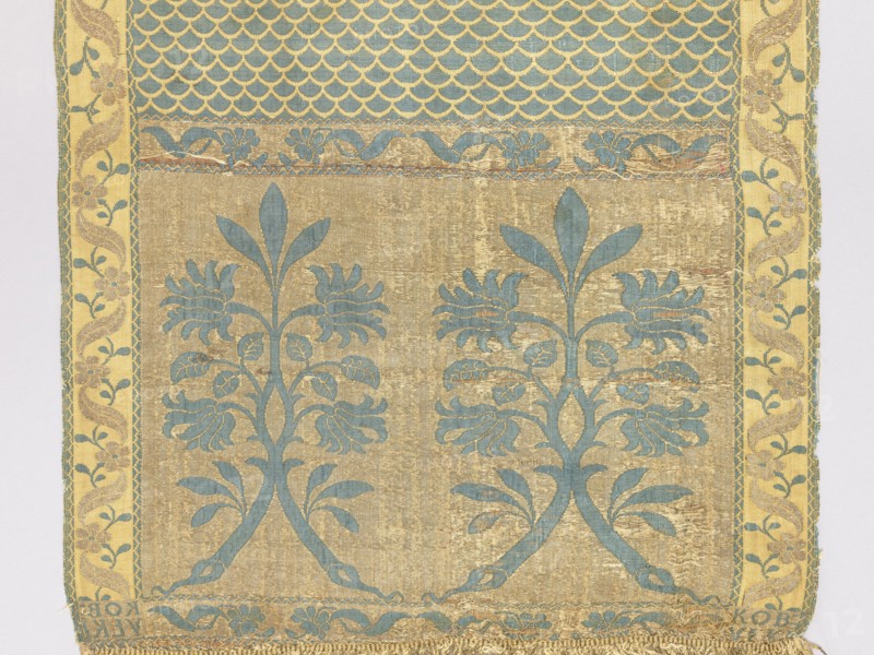 Kontusz belt, Aleksander Unrug Manufactory Kobylka; belt manufacturer; 1781–1787. Solid, double-sided Polish kontusz belt; men’s clothing accessory. Bipartite heads with a motif of two rooted and connected twigs with flowers and leaves forming a bouquet; center with carp-scale motif. Longitudinal border of winding tendrils: leaves with flowers and tendrils of small leaves; crosswise border of flower projections with leaves; additional borders with a snake motif. Sewn-on gold fringe metal and cotton. Ornament: heads—two floral motifs consisting of two branches topped with a common trefoil; center—carp scale motif; borders—vertical and horizontal floral tendril motif intertwined with a wavy ribbon. Colors: obverse—gold background of heads and horizontal borders, blue ornament; straw background of vertical borders, gold and blue ornament; blue background in the center with straw-colored scale pattern. Reverse right. Signed in the heads: KOB:/YLKI. Material/density: approx. 22/27 ×2 silk t...