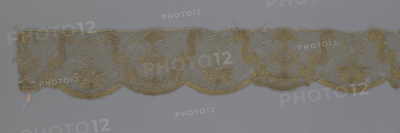 Lace fragment attributed to Kublicki Piottuch or Alfred 1861–1945. Lace strip finished with flat, arched teeth on one side and straight on the other. In the divisions corresponding to the teeth, an alternating flower calyx and a rhombus motif are placed, separated by parenthetical geometric forms. 18th century, France. Material: fabric, lace; width 17.5 cm, length 308 cm., A narrow strip of lace with a scalloped lower edge and a straight upper edge, featuring a repeating embroidered pattern of ornamental shapes and floral motifs; the lace is primarily gold and appears set against a gray background, with areas of fine netting and denser embroidered sections forming curved S-shaped scrolls, diamond-like lattice elements, and stylized flower clusters; the gold thread shows slight variations in sheen and texture, and there are a few loose threads and frayed edges visible along the top and at one end.