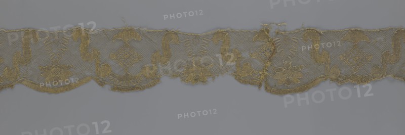 Lace fragment, lace strip finished with flat arched teeth on one side and straight on the other. In the divisions corresponding to the teeth, an alternating flower calyx and a rhombus motif are placed, separated by parenthetical geometric forms. 18th century, France; lace, fabric; width 17.5 cm, length 308 cm., A narrow strip of lace-like fabric with a repeating ornamental pattern rests against a plain background; the fabric is primarily golden with variations of muted gold and tan, and the background is a cool gray. The lace shows a mesh ground with a scalloped lower edge and an upper straight edge, and the decorative motif alternates between curving scroll shapes and floral clusters connected by small geometric lattice elements. The scroll motifs are rounded and sinuous, the floral motifs have layered petal forms, and there are areas where threads are frayed or missing, creating small gaps. The overall texture appears delicate and airy with denser embroidered outlines and open netted...