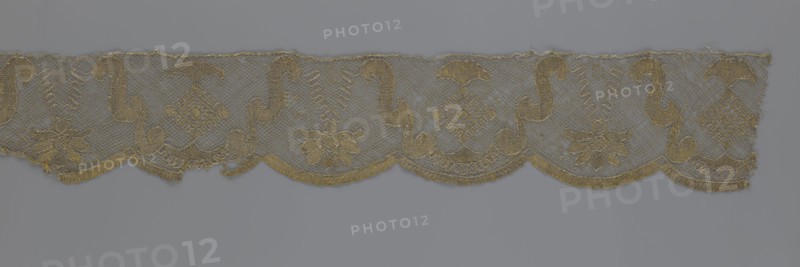 Lace fragment, lace strip finished with flat arched teeth on one side and straight on the other; in the divisions corresponding to the teeth, an alternating flower calyx and a rhombus motif are placed, separated by parenthetical geometric forms. 18th century, France. Materials: lace, fabric. Dimensions: width 17.5 cm, length 308 cm., A horizontal strip of delicate lace with scalloped lower edges and a repeating ornamental pattern featuring curved S-shaped motifs, floral forms, and diamond-like lattice medallions, all rendered in a metallic golden thread against a muted gray background; the lace shows areas of open netting and denser embroidered sections, with fine tonal variations of gold and subtle hints of darker and lighter gray where the background shows through.
