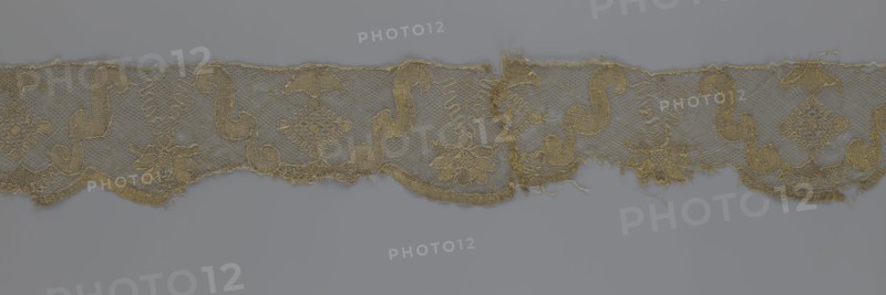 Lace fragment, lace strip finished with flat arched teeth on one side and straight on the other; in the divisions corresponding to the teeth an alternating flower calyx and a rhombus motif are placed, separated by parenthetical geometric forms. 18th century, France. Materials: lace, fabric. Dimensions: width 17.5 cm, length 308 cm., A horizontal strip of delicate lace with a floral and scrolling pattern lies against a flat background; the lace shows a repeating design of flower motifs and sinuous scrolls connected by a fine net ground, with scalloped lower edges and some frayed threads and worn areas; visible colors include shades of muted gold and beige in the lace and a cool gray background.