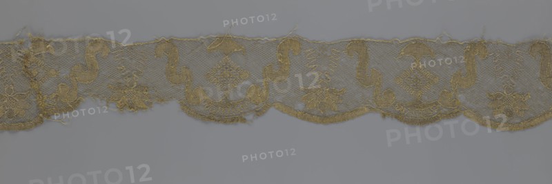 Lace fragment, lace strip finished with flat arched teeth on one side and straight on the other; in the divisions corresponding to the teeth, an alternating flower calyx and rhombus motif are placed, separated by parenthetical geometric forms. 18th century, France; fabric; width 17.5 cm, length 308 cm., A length of delicate lace trim displayed against a neutral background, featuring a repeating scalloped lower edge and a straight upper edge; the lace shows a mesh ground with ornate embroidered motifs including floral shapes, stylized scrolls, and diamond-like elements; the fabric appears slightly frayed in places with loose threads and some irregularities along the edges; visible colors include gold in the embroidery and a muted grayish background.