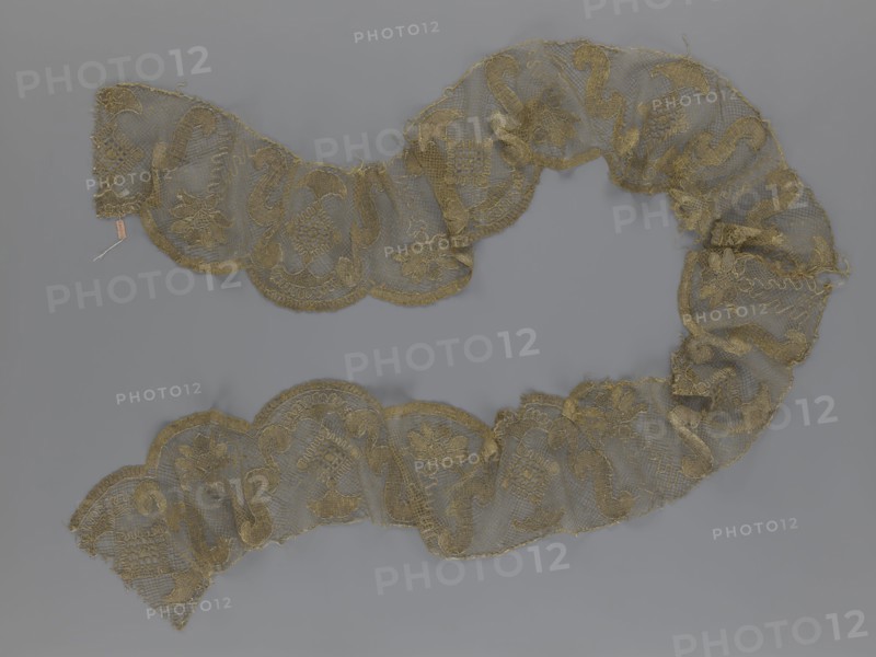 Lace fragment, lace strip finished with flat, arched teeth on one side and straight on the other. In the divisions corresponding to the teeth, an alternating flower calyx and a rhombus motif are placed, separated by parenthetical geometric forms. 18th century, France. Materials: lace, fabric. Dimensions: width 17.5 cm, length 308 cm., A sinuous length of lace arranged in a loose S-shape on a gray background, the lace appearing delicate and semi-transparent with scalloped edges and a repeating floral and scroll motif; the lace is primarily gold in color with variations in tone where threads cluster, and the background is a uniform muted gray.
