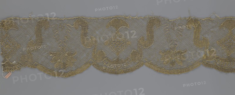 Lace fragment by Kublicki Piottuch, Alfred 1861–1945. The lace strip is finished with flat, arched teeth on one side and straight on the other. In the divisions corresponding to the teeth, an alternating flower calyx and a rhombus motif are placed, separated by parenthetical geometric forms. 18th century, France. Materials: lace, fabric. Width 17.5 cm; length 308 cm., A horizontal strip of ornate lace with a scalloped lower edge displayed against a plain background, showing a repeating embroidered pattern of curving paisley-like shapes, diamond motifs, and floral clusters; the lace has an open netted ground with denser, textured embroidery forming the decorative elements and a slightly frayed top edge with loose threads; visible colors include muted gold thread for the embroidery and a cool gray background.