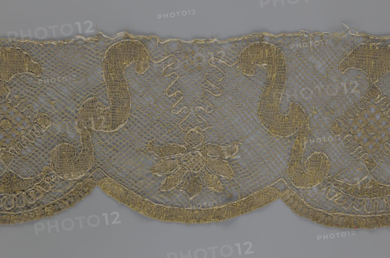 Lace fragment;, The lace strip is finished with flat, arched teeth on one side and straight on the other. In the divisions corresponding to the teeth, an alternating flower calyx and a rhombus motif are placed, separated by parenthetical geometric forms. 18th century, France. Materials: lace, fabric. Dimensions: width 17.5 cm, length 308 cm., The image shows a decorative textile strip with an open net-like background in gray and gold tones, featuring ornate gold embroidered motifs including curved scroll shapes and a central floral element; the lower edge has a scalloped border with dense gold embroidery, and there are a few small loose threads and irregularities along the top edge.