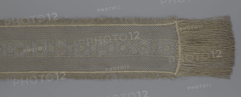 A so-called Saxon lace belt, lace, belts, clothing accessories, men's clothing, fashion, provenance unknown. The central part is a continuous belt with a geometric ornament; among the motifs there is a circle containing an hourglass. The side belts of similar width are permanently connected. The entire belt is trimmed with fringe. 17th/18th century, Poland, lace, clothing accessories, height 270 cm, width 25 cm., A horizontal strip of woven fabric with fringed edges running along the top and bottom and a longer fringe at the right end, the textile shows a central patterned band with an open lattice structure flanked by areas of tighter net-like weave; the material appears in shades of beige and gray with variations of light tan and muted brown in the threads, set against a smooth gray background.