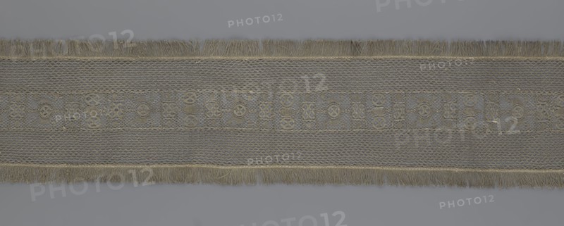A so-called Saxon lace belt, creator, lace belt for men's clothing; central continuous belt with a geometric ornament including a circle containing an hourglass; side belts of similar width permanently connected; entire belt trimmed with fringe; 17th–18th century, Poland; lace clothing accessory; height 270 cm, width 25 cm., A long horizontal strip of woven fabric with fringed edges, showing a repeated decorative pattern along its center; the piece displays a textured mesh background with a band of geometric and circular motifs, and fringes at the top and bottom. The visible colors are shades of gray, muted beige, and pale gold.