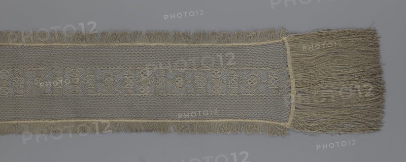 A so-called Saxon lace belt, lace belt accessory for men's clothing. The central part is a continuous belt with a geometric ornament, including a motif of a circle containing an hourglass. The side belts of similar width are permanently connected. The entire belt is trimmed with fringe. 17th/18th century, Poland; lace clothing accessory; height 270 cm, width 25 cm., A long, narrow piece of woven fabric with fringe on both short ends and along one long edge, displayed on a gray background; the fabric shows an open, net-like weave in a beige or tan color with a central decorative band featuring a repeating geometric motif woven in the same beige or tan hue, and a denser woven border separating the decorative band from the netting; a wide tassel-like fringe of loose beige or tan threads extends from one short end.