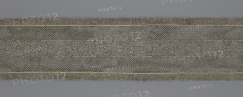 A so-called Saxon lace belt, lace belt accessory for men's clothing. The central part is a continuous belt with a geometric ornament; among the motifs is a circle containing an hourglass. The side belts of similar width are permanently connected. The entire belt is trimmed with fringe. 17th–18th century, Poland. Lace, clothing accessory; height 270 cm, width 25 cm., A long horizontal textile band with fringed edges runs across a neutral background; the band shows a central decorative strip flanked by textured panels, all in muted tones of beige, tan, gray, and hints of olive. The fringes at the top and bottom edges are a mix of beige and tan threads. The central strip contains a repeating mosaic of small geometric motifs created by raised stitching in beige and olive against a mottled gray-beige ground. Adjacent panels on either side of the central strip display a fine net-like or lattice texture in pale gray and beige, with subtle variations in thread thickness creating a slightly thr...