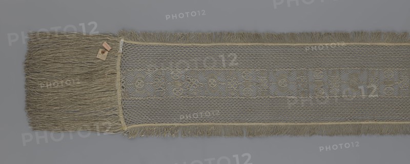 A so-called Saxon lace belt, lace, belts, clothing accessories, men's clothing, clothing, fashion, deposit provenance. The central part is a continuous belt with a geometric ornament; among the motifs is a circle containing an hourglass. The side belts of similar width are permanently connected. The entire belt is trimmed with fringe. 17th/18th century, Poland; lace, clothing accessories, clothing; height 270 cm, width 25 cm, A long, narrow woven textile with a central decorative band and fringed edges, showing shades of beige, tan, gray, and muted gold; one end displays a dense group of long, loose fringe strands, while the main body features an open, lacy mesh pattern with a repeating geometric motif and shorter fringe along the sides, all set against a plain gray background.