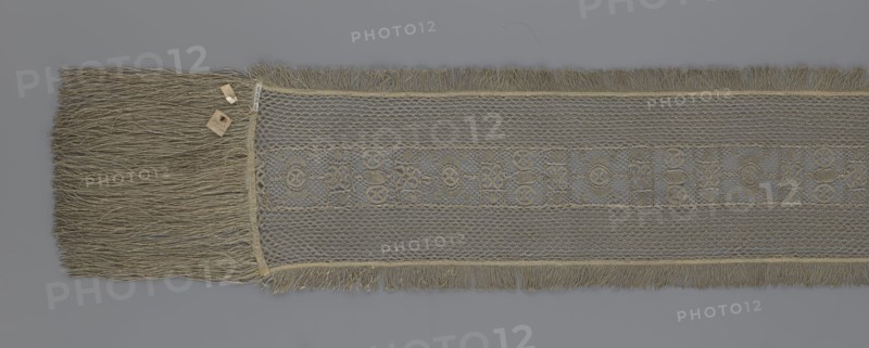A so-called Saxon lace belt, lace, belts, clothing accessories, men's clothing, clothing, fashion, deposit provenance. The central part is a continuous belt with a geometric ornament; among the motifs is a circle containing an hourglass. The side belts of similar width are permanently connected. The entire belt is trimmed with fringe. 17th/18th century, Poland. Lace, clothing accessories, clothing. Height 270 cm, width 25 cm., A long woven textile strip lies flat against a gray background, showing a central band with a repeating geometric openwork pattern flanked by finer mesh panels and fringed edges; the textile and its fringe appear in shades of tan, beige, and muted gold, with the mesh areas showing slightly darker and lighter variations of those colors, and a small cluster of tan-colored tags or paper pieces rests near one end on top of the fringe.