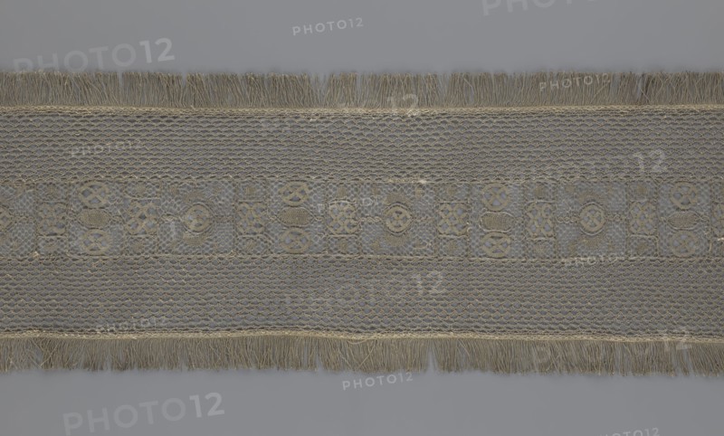 A so-called Saxon lace belt, lace belt and clothing accessory for men's clothing, decorated with a continuous central belt featuring a geometric ornament including a circle containing an hourglass. Side belts of similar width are permanently connected. Entire belt trimmed with fringe. 17th–18th century, Poland. Materials: lace. Dimensions: height 270 cm, width 25 cm., A horizontal band of woven fabric displayed against a gray background, featuring a central decorative strip with an openwork pattern of interlocking shapes in beige and tan tones, flanked above and below by sections of netlike mesh in similar beige and light brown hues, and finished at the top and bottom edges with fringe strands in beige and light tan.