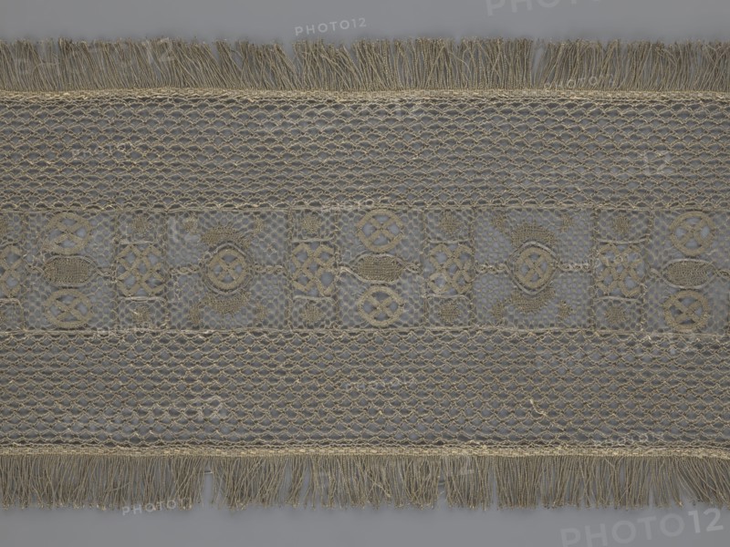 A so-called Saxon lace belt, creator, lace belt men's clothing accessory. The central part is a continuous belt with a geometric ornament, including a circle containing an hourglass motif. Side belts of similar width are permanently connected. The entire belt is trimmed with fringe. 17th–18th century, Poland. Lace; height 270 cm, width 25 cm., A rectangular piece of lace fabric laid flat with a repeating horizontal pattern, featuring a central band with ornate geometric motifs and surrounding bands of open net-like stitching; fringed edges run along the top and bottom. The visible colors are shades of beige and tan for the lace against a muted gray background, with subtle variations where the threads overlap. The lace shows textured braided borders, hexagonal and diamond openwork, and circular and interlaced decorative shapes in the central area, all in the same beige-tan tones. The fringe consists of loose hanging threads in beige and tan, and the overall surface displays fine threadw...
