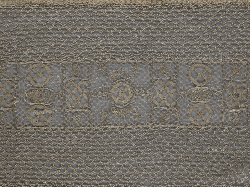 A so-called Saxon lace belt, lace belt/men's clothing accessory. The central part is a continuous belt with a geometric ornament; among the motifs is a circle containing an hourglass. Side belts of similar width are permanently connected. The entire belt is trimmed with fringe. 17th/18th century, Poland. Materials: lace. Dimensions: height 270 cm, width 25 cm., A close-up of a textured fabric with an open, net-like weave in beige and gray tones, featuring a central horizontal band with a decorative pattern of embroidered geometric motifs and oval shapes; the surrounding areas show a repeating diamond mesh with braided threads, and the overall palette includes shades of beige, tan, light brown, and muted gray.
