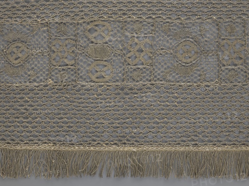A so-called Saxon lace belt, creator unknown, lace belt for men's clothing, clothing accessory; central continuous belt with a geometric ornament including a circle containing an hourglass; side belts of similar width permanently connected; entire belt trimmed with fringe; 17th–18th century, Poland; lace; height 270 cm, width 25 cm., A close-up view of a textile featuring a repeating openwork pattern in beige and gray tones, with a top band of decorative embroidered motifs set against a gray background, a middle section of a netlike lattice, and a lower fringe of loose threads; the embroidery shows several circular and diamond-shaped stitched elements in beige, the net section displays a repeating diamond mesh also in beige over gray, and the bottom fringe consists of a group of hanging beige threads.